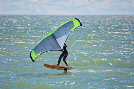Man is wing foiling using handheld inflatable wing and hydrofoil surfboard in sea in summer. Rider on a wind wing board, surfs the waves.の写真素材