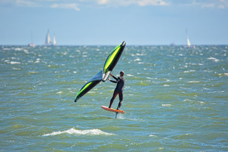 Man is wing foiling using handheld inflatable wing and hydrofoil surfboard in sea in summer. Rider...