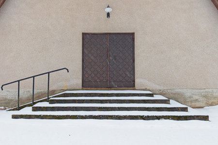 Old weatherd wooden double  doors on a stone building with steps and handrail in winter with snow on the ground.の写真素材