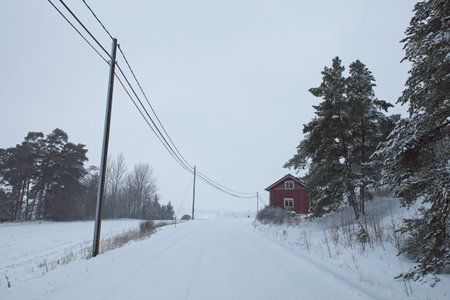 Snow covered road with red painted house in cloudy winter weather.の写真素材