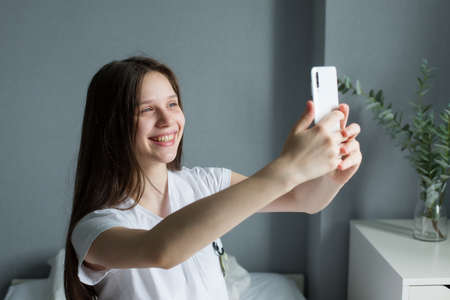 Teenage girl is sitting in her pajamas on her bed, takes a selfie on her smartphone.の写真素材