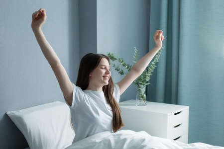 Happy teenage girl meets a new day in a beautiful room. A beautiful teenager wakes up in the morning, stretches in bed, bright light on her from a large window.の写真素材