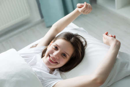 Portrait Of A Beautiful Young Brunette Teenager Lying in Her Bed and Smiling at The Camera, Her Face Illuminated By The Warm morning sun. Sweet and Warm.の写真素材