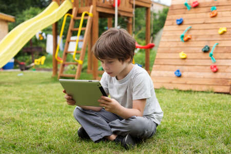 Online training, online games, a boy with a tablet is sitting on the green grass on the playground.の写真素材