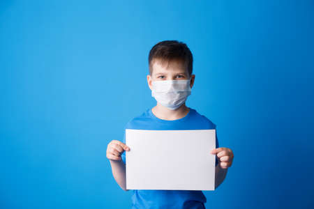 Boy in a blue T-shirt on a blue background, holding a white cardboard on a blue background, a schoolboy smiles and holds an advertising sheet, a place to copy.の写真素材