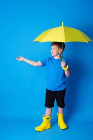a boy in a blue T-shirt, yellow boots on a blue background holds a yellow umbrella, a schoolboy smiles, place to copy.の写真素材