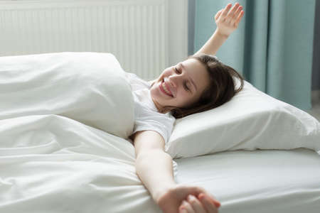A beautiful teenager wakes up in the morning, stretches in bed, bright light on her from a large...