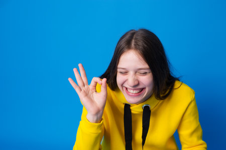a girl with long hair in a yellow sweatshirt smiles with her eyes closed and shows an okey gesture...
