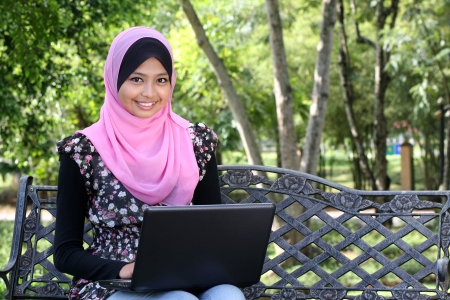 Beautiful muslim woman using laptop while sitting relaxed on bench at summer park の写真素材