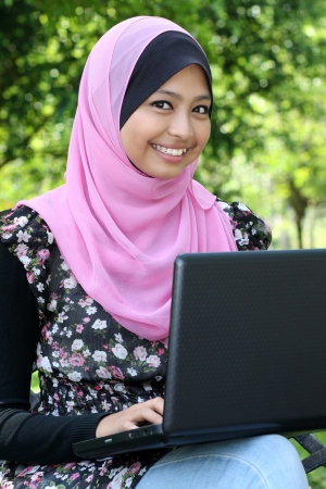 Beautiful muslim woman using laptop while sitting relaxed on bench at summer park の写真素材