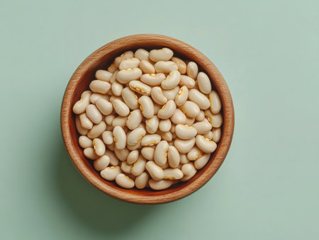 a top view of a wooden bowl packed with white beans, sitting against a light green backdrop. the image highlights the natural texture of the beans.の素材