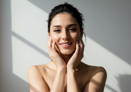 A beautiful woman smiles while gently touching her face with her hands. The image highlights her radiant skin and natural beauty, suggesting a focus on skincare, wellness, or cosmetic treatments. The soft lighting enhances the overall aesthetic, creating a serene and healthy impression.の素材