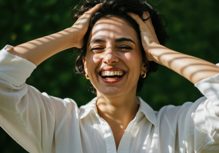A smiling woman is shown with her hands in her curly hair. She is wearing a white shirt and gold earrings. The image conveys happiness and joy, with a blurred green background.の素材