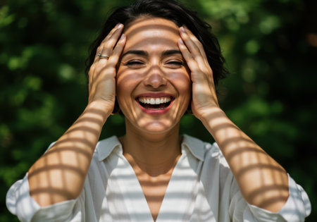 A smiling woman is bathed in sunlight, creating interesting shadow patterns across her face. She has her hands near her head and is wearing a light-colored shirt. The background is filled with green foliage, suggesting an outdoor setting.の素材