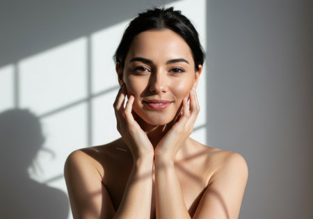 A portrait of a beautiful woman with her hands gently touching her face. The image showcases a clean, fresh look with natural light and shadows enhancing her features. The focus is on beauty, wellness, and healthy skin.の素材