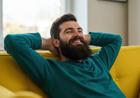 A happy, bearded man with his hands behind his head, looking up and smiling while relaxing on a comfortable yellow couch in a home setting.の素材