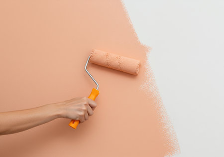 A close-up shot shows a hand holding an orange paint roller, applying a fresh coat of peach-colored paint to a white wall, transforming the space.の素材