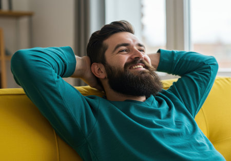 A bearded man with a warm smile rests comfortably on a vibrant yellow sofa, his hands clasped behind his head, enjoying a moment of relaxation indoors.の素材