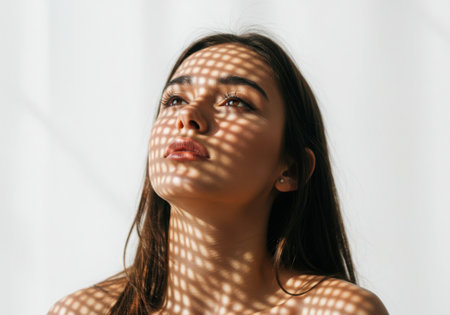 A close-up portrait of a young woman looking upwards, her face illuminated by sunlight casting a grid-like shadow pattern across her features.の素材