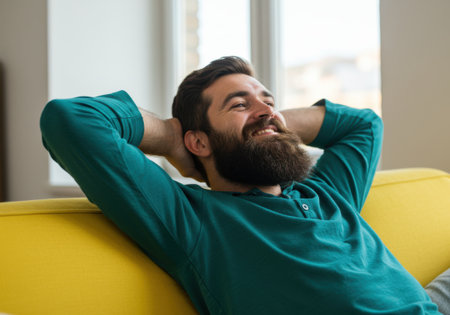 A content bearded man with his hands behind his head, smiling as he relaxes on a comfortable yellow couch in a bright, indoor setting.の素材
