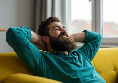 A bearded man with his arms behind his head smiles contentedly while relaxing on a bright yellow sofa indoors, enjoying a moment of peace.の素材