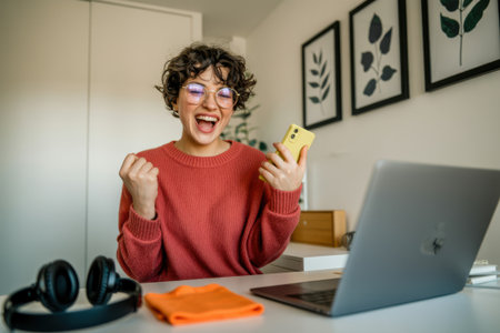 A joyful woman with curly hair cheers enthusiastically in a well-lit home office. She's holding cash, her laptop open, and headphones nearby, signifying a moment of triumph.の素材