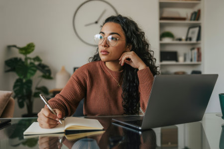 A focused young woman with curly hair and glasses is diligently working on her laptop and taking notes in a notebook. The scene evokes a productive and studious atmosphere in a comfortable home environment.の素材