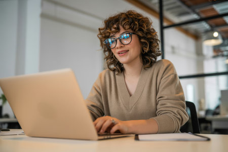 A smiling young woman with glasses and curly hair focuses on her laptop screen in a contemporary, well-lit office. She appears engaged in her work, surrounded by the professional environment.の素材