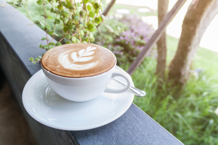 Closeup of white cup with hot latte art on top wooden board in garden with lighting on top right sideの写真素材