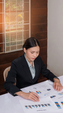 Young asian businesswoman sitting at workplace and reading paper in officeの写真素材
