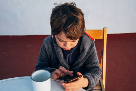 Child with a smartphone drinking coffee at breakfast while using his mobile phone to call, chat and...