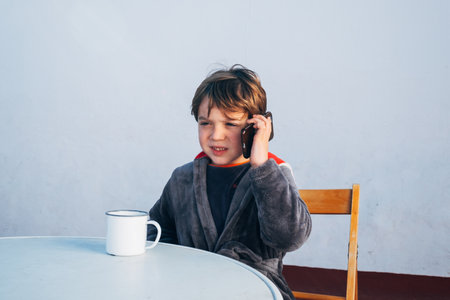 Child with a smartphone drinking coffee at breakfast while using his mobile phone to call, chat and read the newsの写真素材