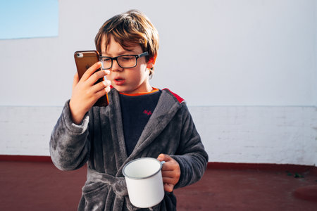 Child with a smartphone drinking coffee at breakfast while using his mobile phone to call, chat and...