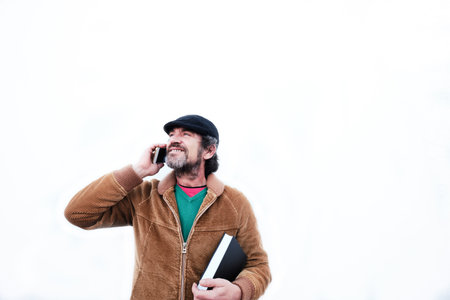 Man in a cap and brown jacket with a book in his hand while talking on the mobile phoneの写真素材
