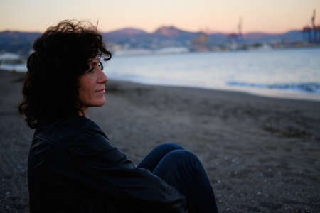 Adult woman sitting on the beach looking at the sea during sunset one winter dayの写真素材