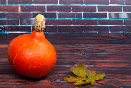 The photo shows an orange Hokkaido pumpkin on a wooden table with a leaf in front of a wallの写真素材