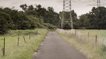 A cyclist passes a high voltage pylon on a road in the meadows.の写真素材