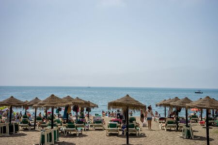 Malaga, Spain - June 24: People basking in the sun at Malagueta beach under beach huts with the ocean and a boat in the background in Malaga, Spain, Europe on a cloudy dayのeditorial素材