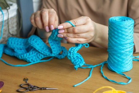 Hands of a young woman knit crochet from blue yarn on a light wooden surface. Start a new bag of yarn. Selective soft focus.の写真素材