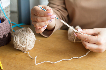 Hands of a young woman knit crochet from beigeyarn on a light wooden surface. Start a new bag of...