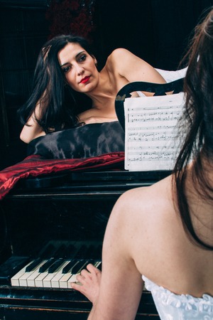 A young woman in vintage lingerie is lying on the piano. Her girlfriend plays music on the piano by the notes. Added a small grain, imitation of film photography.の写真素材