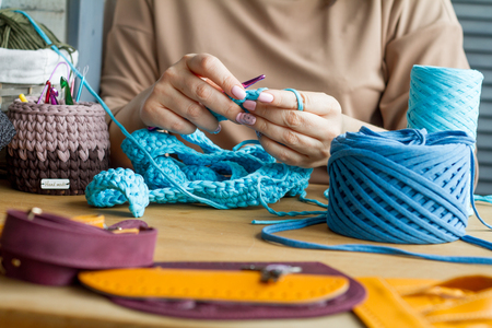 Hands of a young woman knit crochet from blue yarn on a light wooden surface. Start a new bag of yarn. Selective soft focus.の写真素材