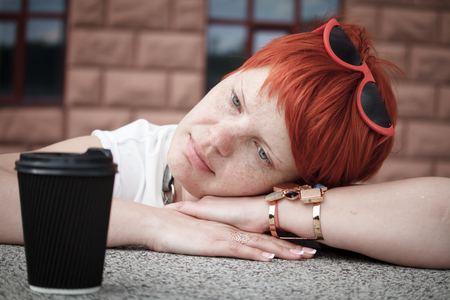 Close-up of a young woman with short red hair walking around the city with a couple of cups of coffee, free space for textの写真素材