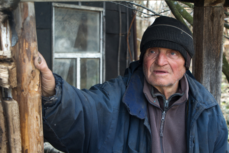 An old man in sloppy clothes stands on the threshold of his own ruined house and looks into the distance, loneliness in the village, life beyond povertyの写真素材