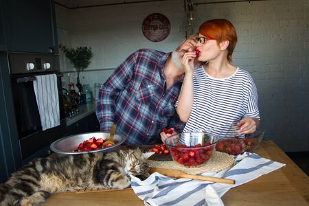 Adult couple man and woman peel and cut strawberries for strawberry jam, feed each other, laugh and have fun, the Maine Coon kitten sleeps on the kitchen tableの写真素材