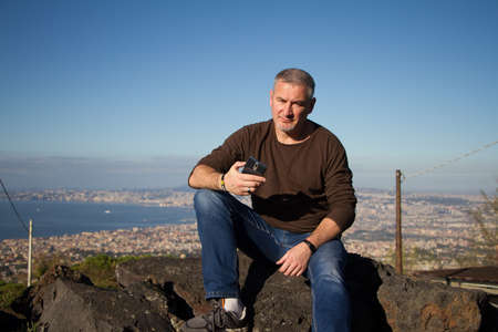 Adult gray-haired man with a beard sits on top of Mount Vesuvius with a view of Naples at sunrise, copyspaceの写真素材