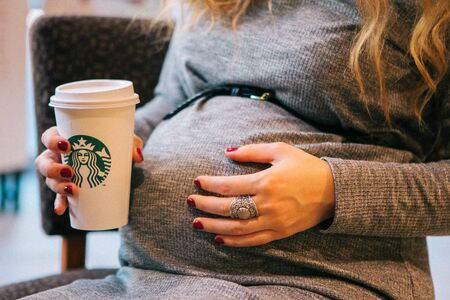 Geneva, Switzerland - October 15, 2017: Close-up belly of a pregnant woman in a gray dress that holds a glass of starbucks, selective focusのeditorial素材