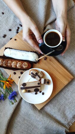 Top view on female hands with summer flowers cornflower and rutbeckia near cup of black coffee and served eclairs, the concept of a pleasant breakfast, selective focusの写真素材
