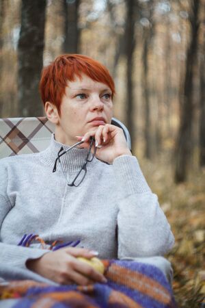 Red-haired woman sitting in a folding deck chair sheltered by a bright woolen plaid on blurred background of an autumn forest, selective focusの写真素材