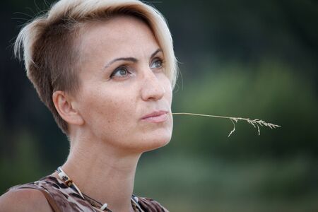 Close up of a middle-aged blond woman with a spikelet in her mouth looking thoughtfully to the side...